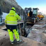 Grävschakt som är täck med körplåt. Byggarbetare i förgrunden och grävmaskin i bakgrunden